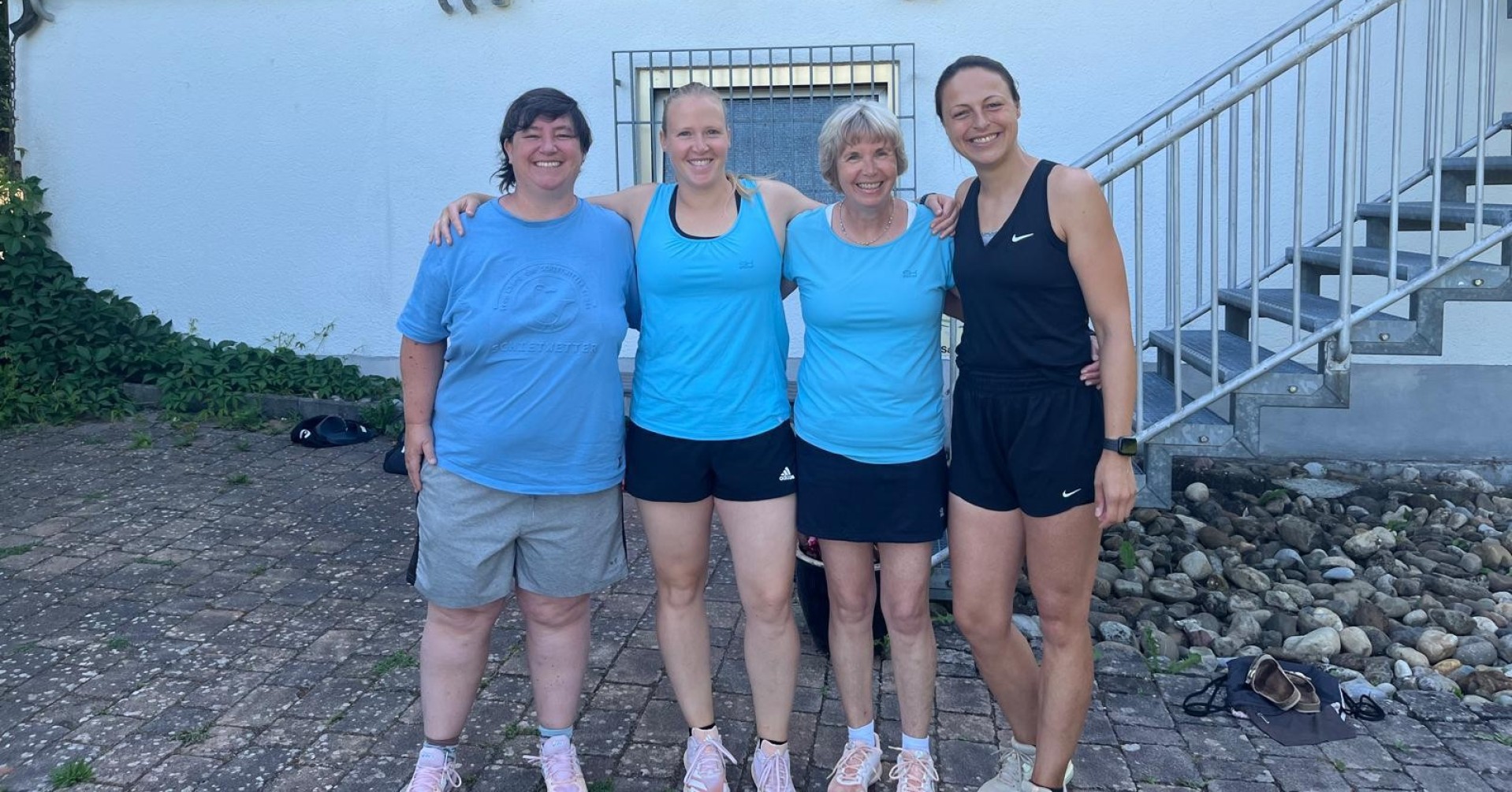 Vier Frauen in Sportkleidung stehen gemeinsam vor einem Vereinsgebäude und lächeln in die Kamera. Sommerliche Szene, Gruppenfoto nach dem Sport.