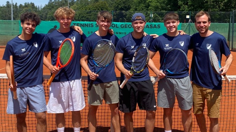 Junge Tennisspieler stehen mit Schlägern auf dem Tennisplatz Vier junge Männer mit Tennisschlägern stehen auf einem Tennisplatz vor einem Netz und lächeln in die Kamera.