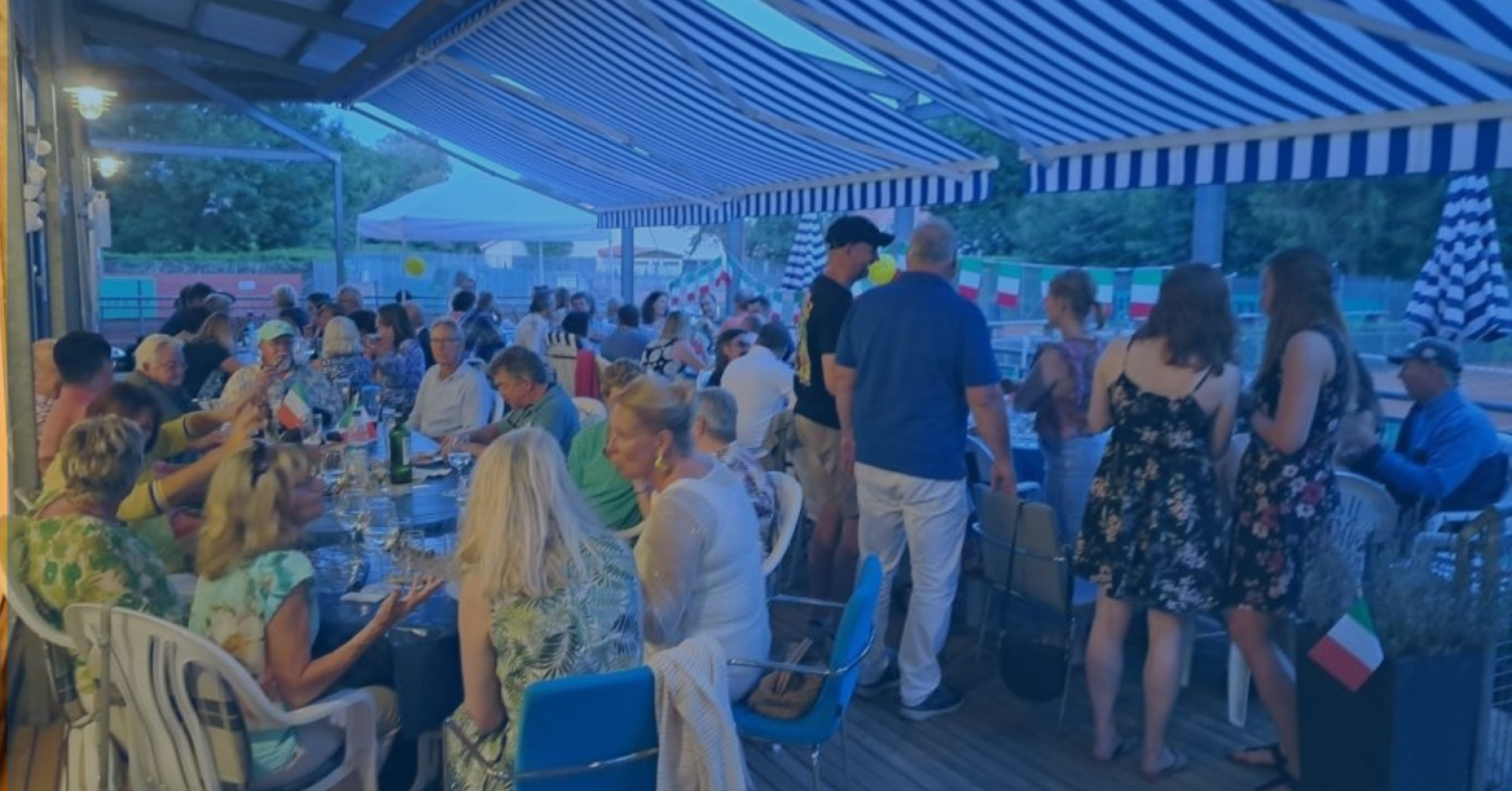 Sommerfest mit vielen Gästen unter blau-weiß gestreiftem Sonnendach, gesellige Atmosphäre, Veranstaltung mit Dekoration und internationalen Flaggen