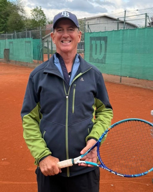 Mann mit Tennisschläger auf Sandplatz, lächelnd, sportliche Kleidung, Tennisplatz im Hintergrund
