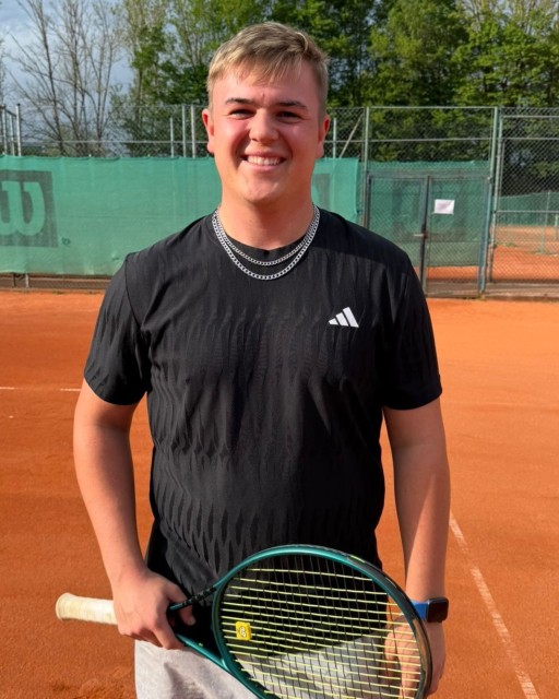 Junger Mann mit Tennisschläger auf Sandplatz, lachend, im schwarzen Sportshirt