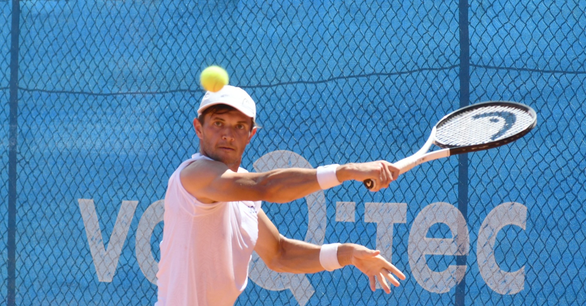 Ein Tennisspieler in weißem Shirt beim Vorhandschlag auf dem Tennisplatz mit blauem Hintergrund.