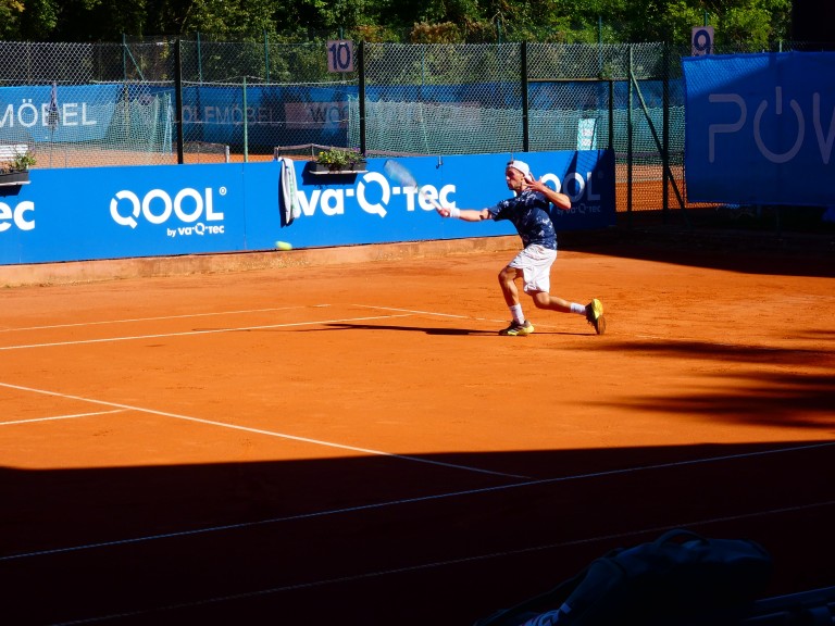 Tennisspieler auf Sandplatz mit QOOL und va-Q-tec Sponsorenbande Tennisspieler auf Sandplatz beim Schlag, Sponsorenbande QOOL und va-Q-tec im Hintergrund