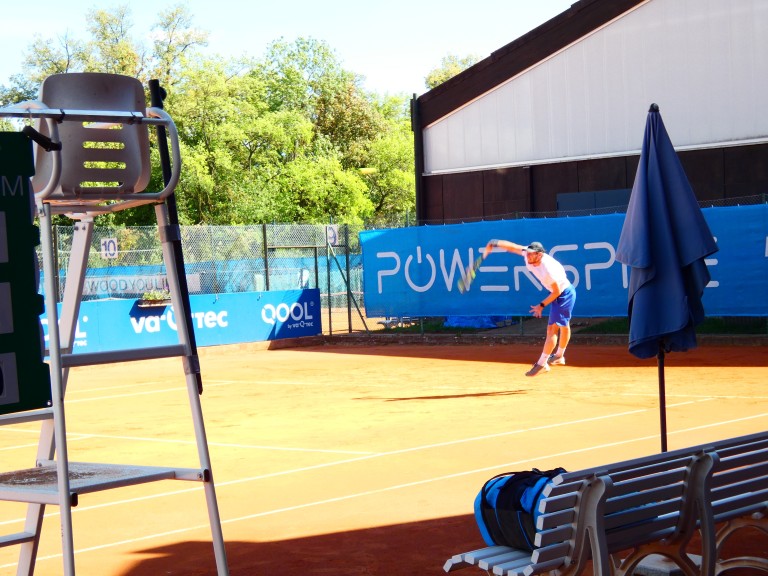 Tennisspieler beim Aufschlag auf Sandplatz unter blauem Himmel Tennisspieler beim Aufschlag auf Sandplatz, sonniger Tag, Tennisplatz mit Werbebannern