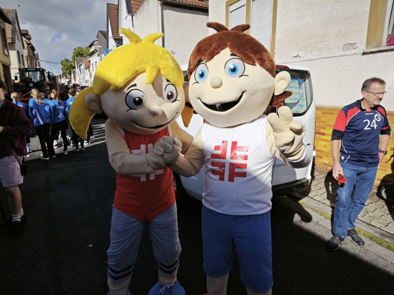 Zwei Maskottchen in Sportkleidung mit Turnfest-Logo bei einem Straßenfest oder Sportevent