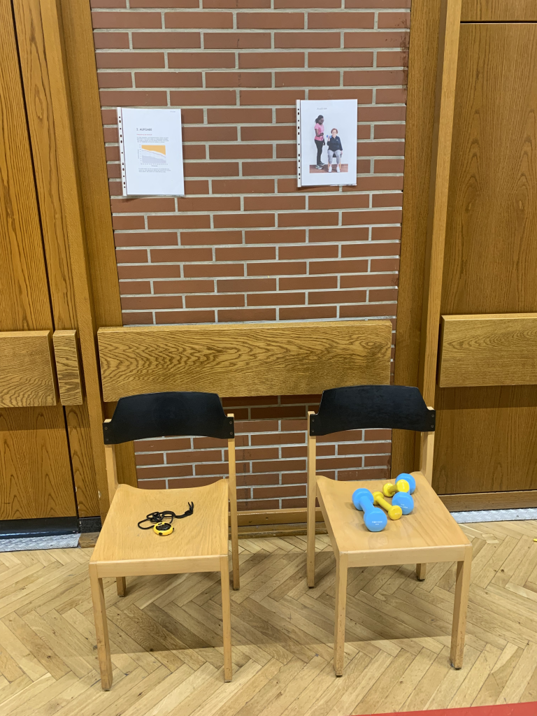 Zwei Stühle mit Fitnessgeräten wie Hanteln und Stoppuhr vor einer Turnhallenwand, vermutlich für einen AFT-Test oder Fitness-Test vorbereitet.