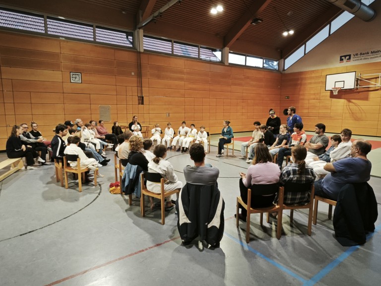 Gruppe von Kindern und Erwachsenen in einer Sporthalle bei einer Besprechung, einige Kinder tragen weiße Judoanzüge