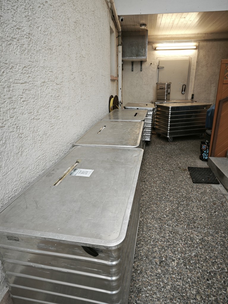Mehrere große Metallboxen in einem Hinterhof oder Lagerbereich, gestapelt auf einem gepflasterten Boden neben einer weißen Wand.