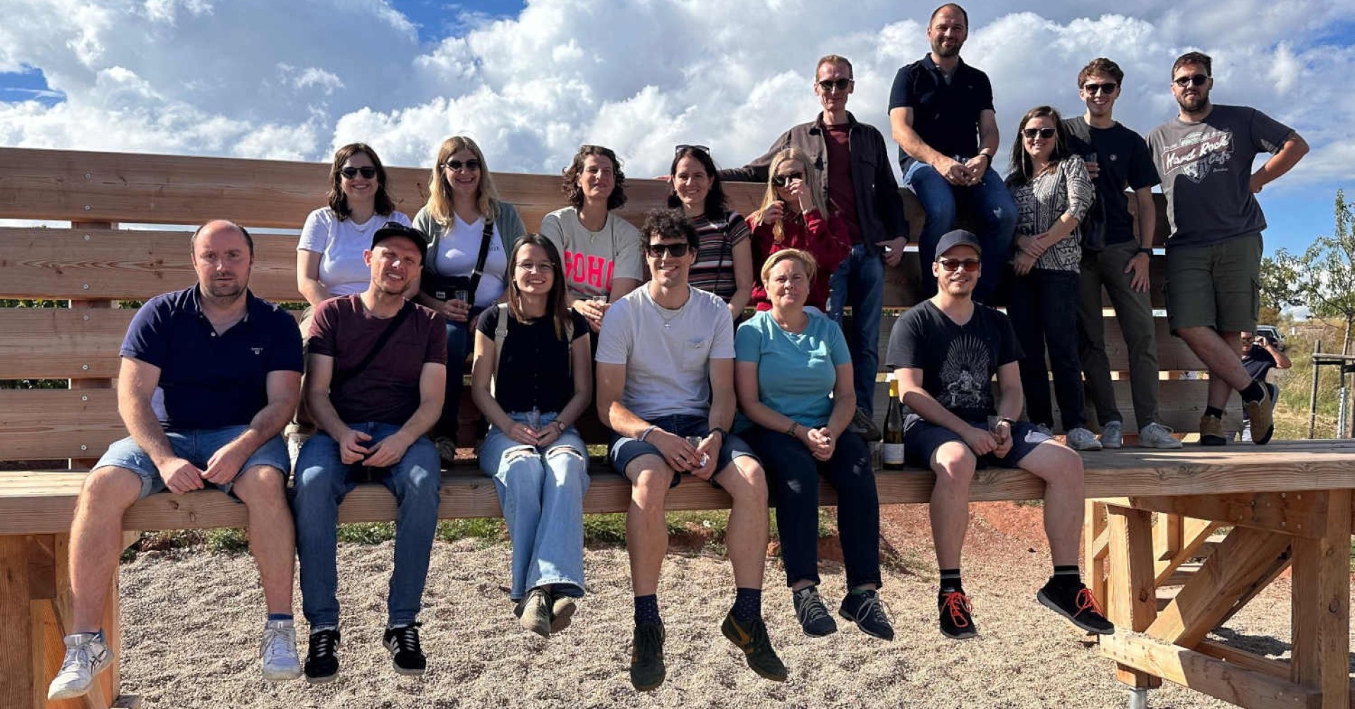 Gruppe von Menschen bei einer Sommeraktivität im Freien auf einer Holzbank unter blauem Himmel