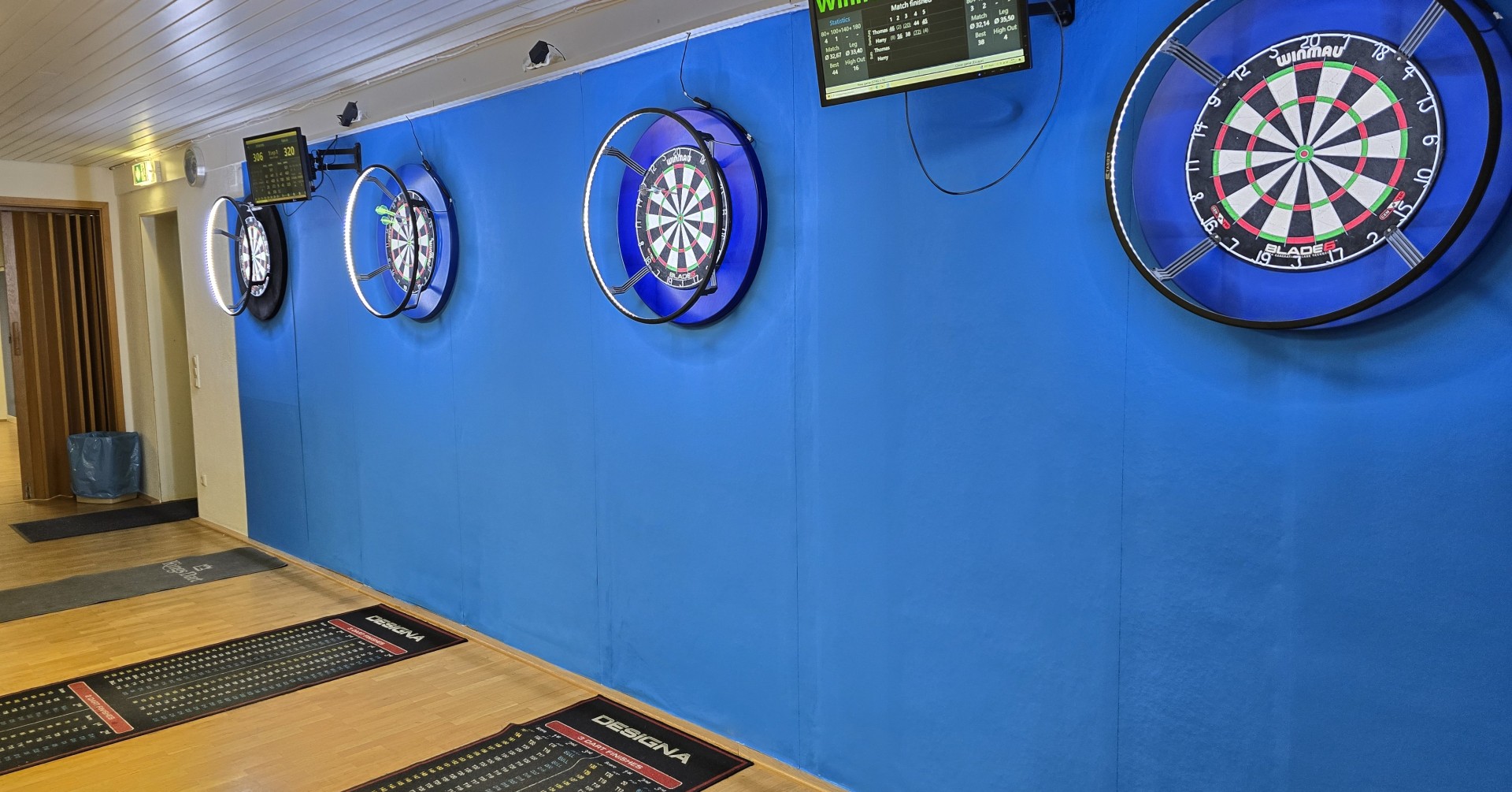 Moderner Darts-Trainingsraum mit Dartboards, elektronischen Anzeigetafeln und Designa-Matten Dartboards und elektronische Anzeigetafeln in einem modernen Darts-Trainingsraum mit blauer Wand und Designa-Matten.