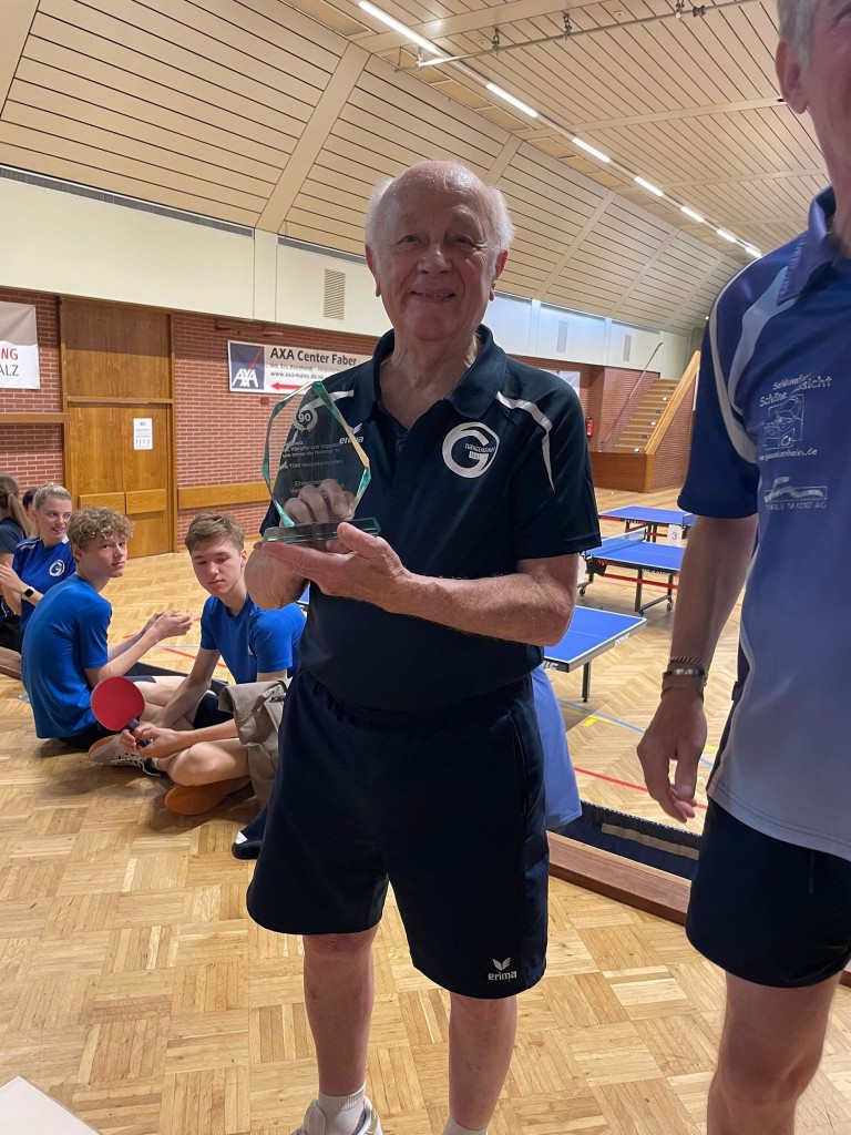Tischtennis-Auszeichnung für Senior in Sporthalle am 21.10.2025 Senior erhält Tischtennis-Auszeichnung in Sporthalle mit Jugendlichen im Hintergrund