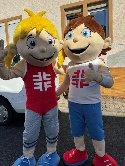 Zwei Maskottchen mit Sportkleidung und Turnfest-Logo stehen nebeneinander und lächeln in die Kamera