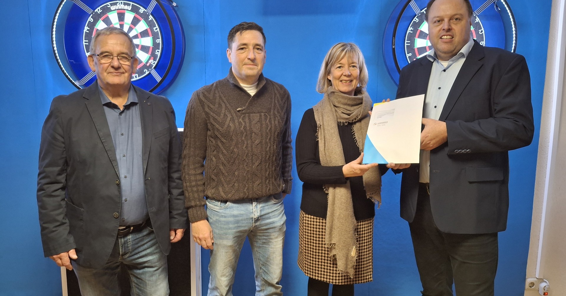 Vier Personen stehen in einem Raum mit blauer Wand, zwei Dartscheiben und einem Bildschirm, eine Person überreicht ein Dokument.