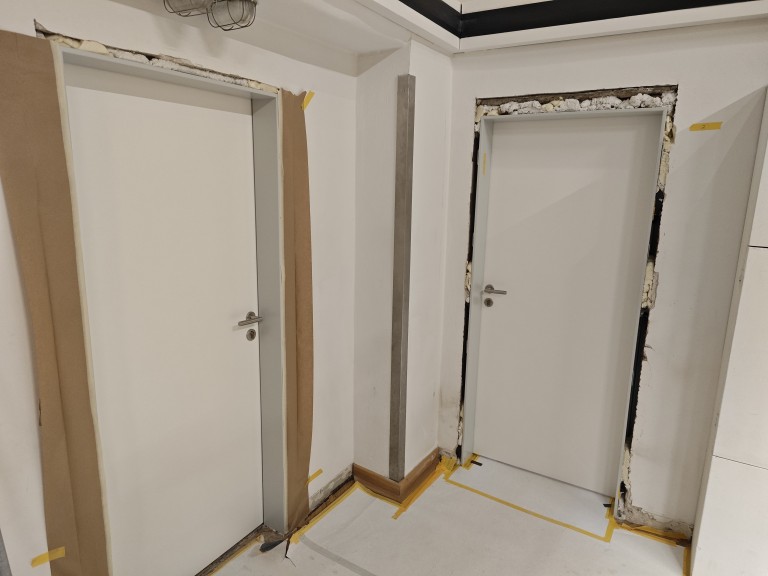 Zwei weiße Türen in einem Innenraum während Renovierungsarbeiten, mit abgekleistertem Boden und unvollständigen Türrahmen.