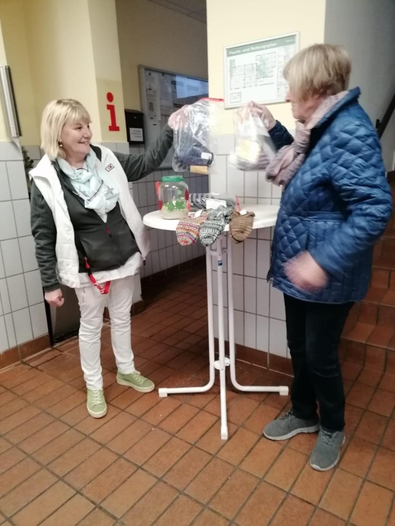 Zwei Frauen tauschen an einem Stehtisch warme Kleidung und Alltagsgegenstände, Symbol für Armut und Gesundheit