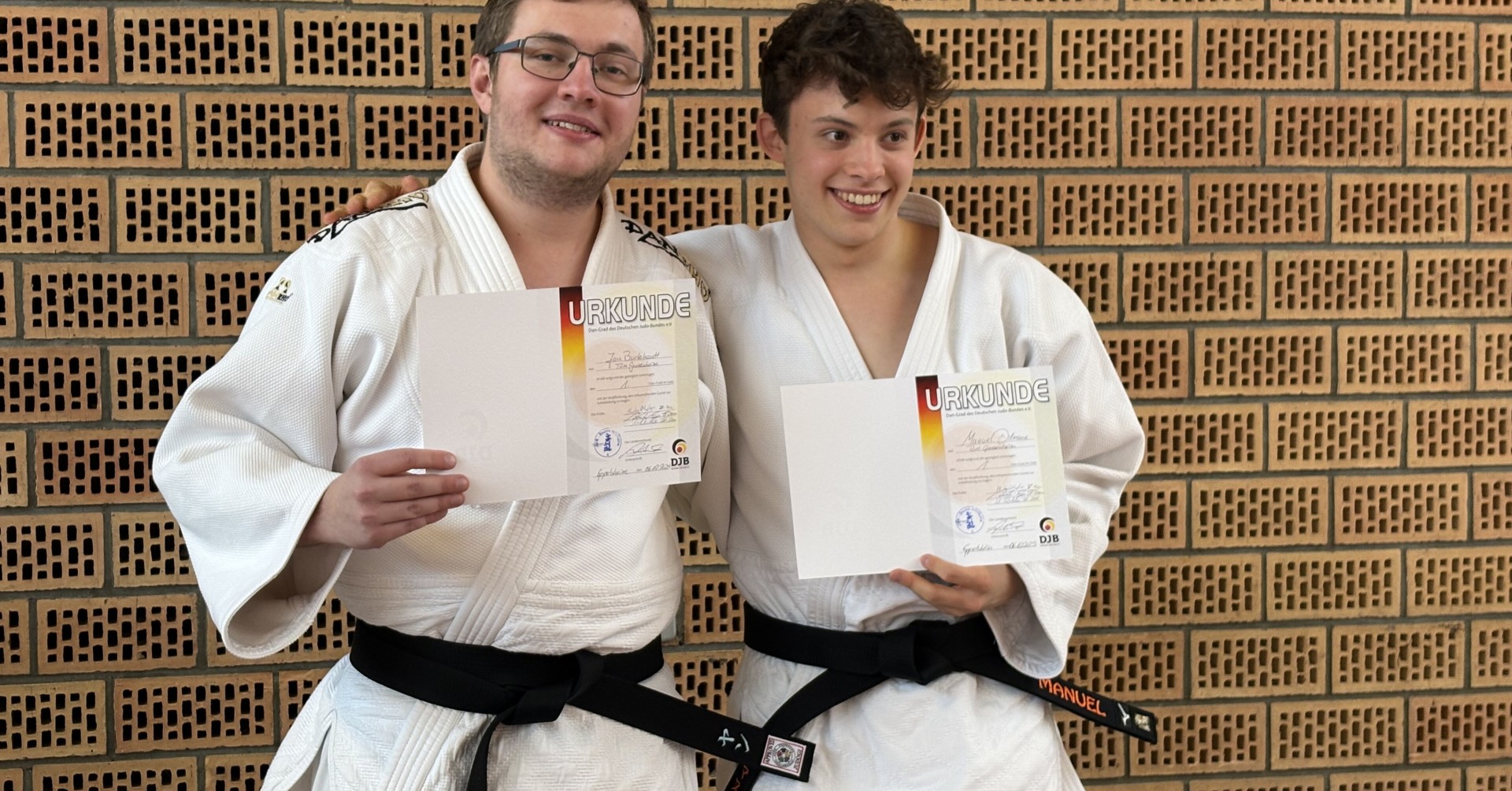 Zwei Judoka mit schwarzem Gürtel halten Urkunden vor einer Backsteinwand.