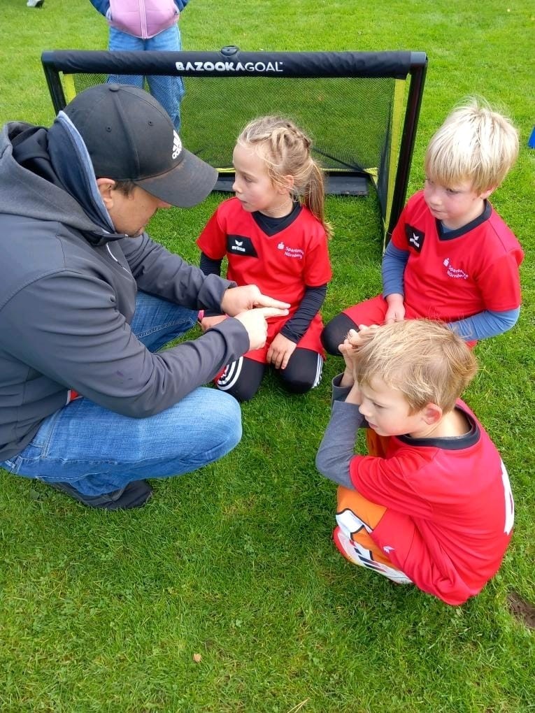 Trainer gibt Kindern im roten Trikot Anweisungen beim Fußballtraining auf dem Sportplatz