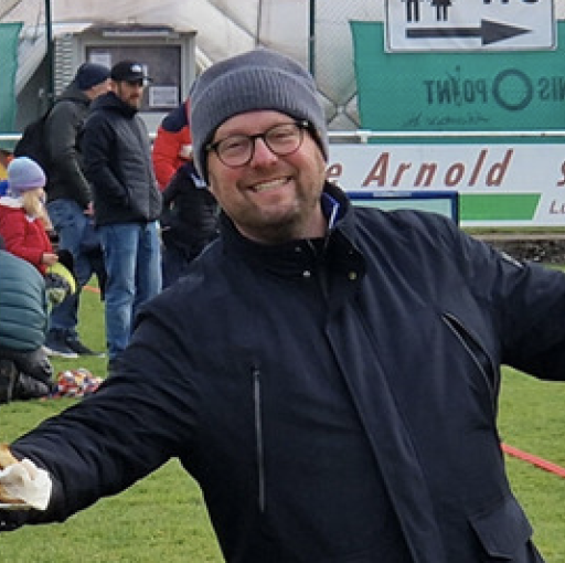 Person mit dunkler Jacke und Mütze auf einem Sportplatz, im Hintergrund Zuschauer und Werbebanner