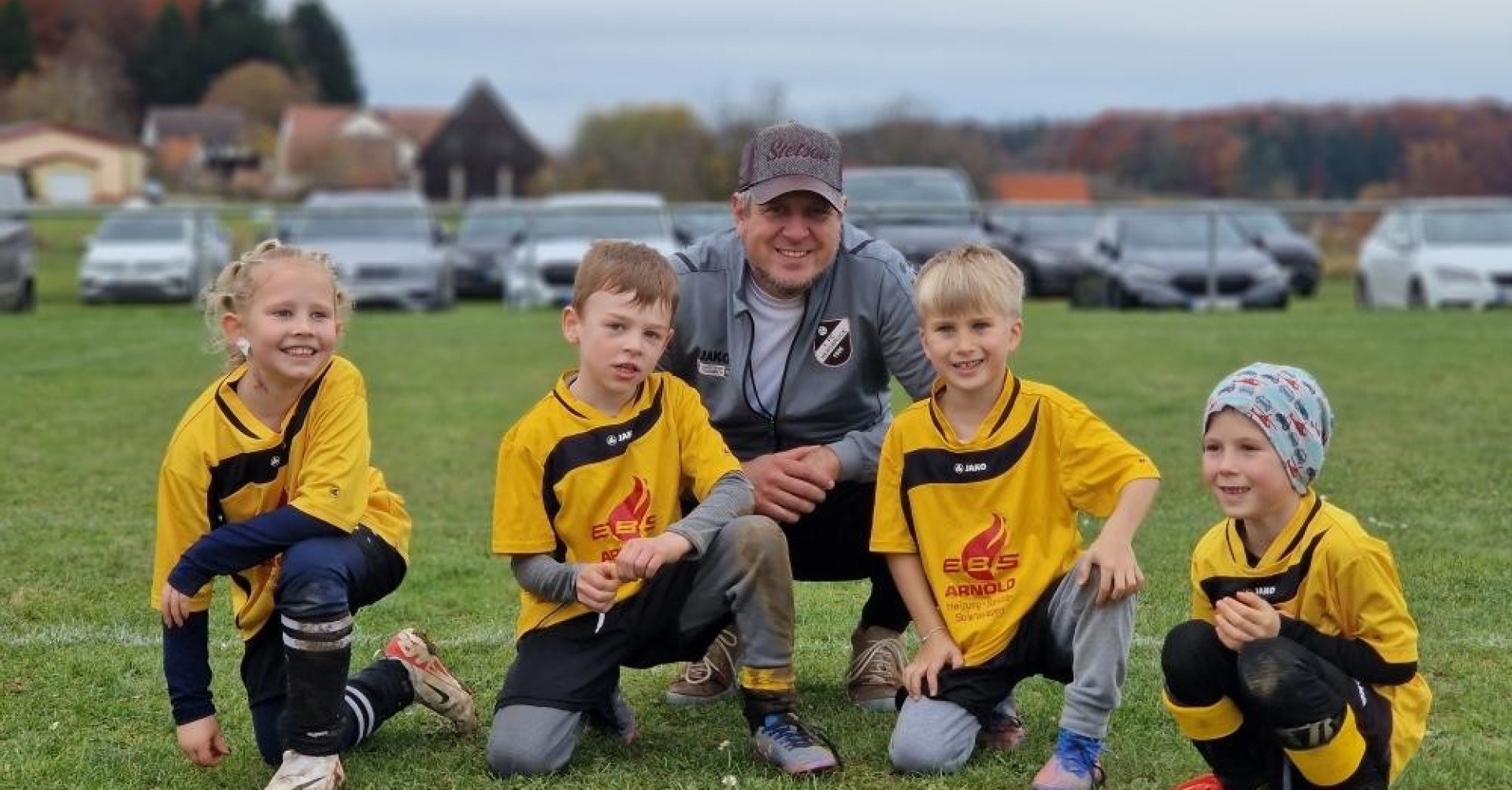 Kinderfußballmannschaft mit Trainer auf dem Sportplatz, gelbe Trikots mit EBS Arnold Logo