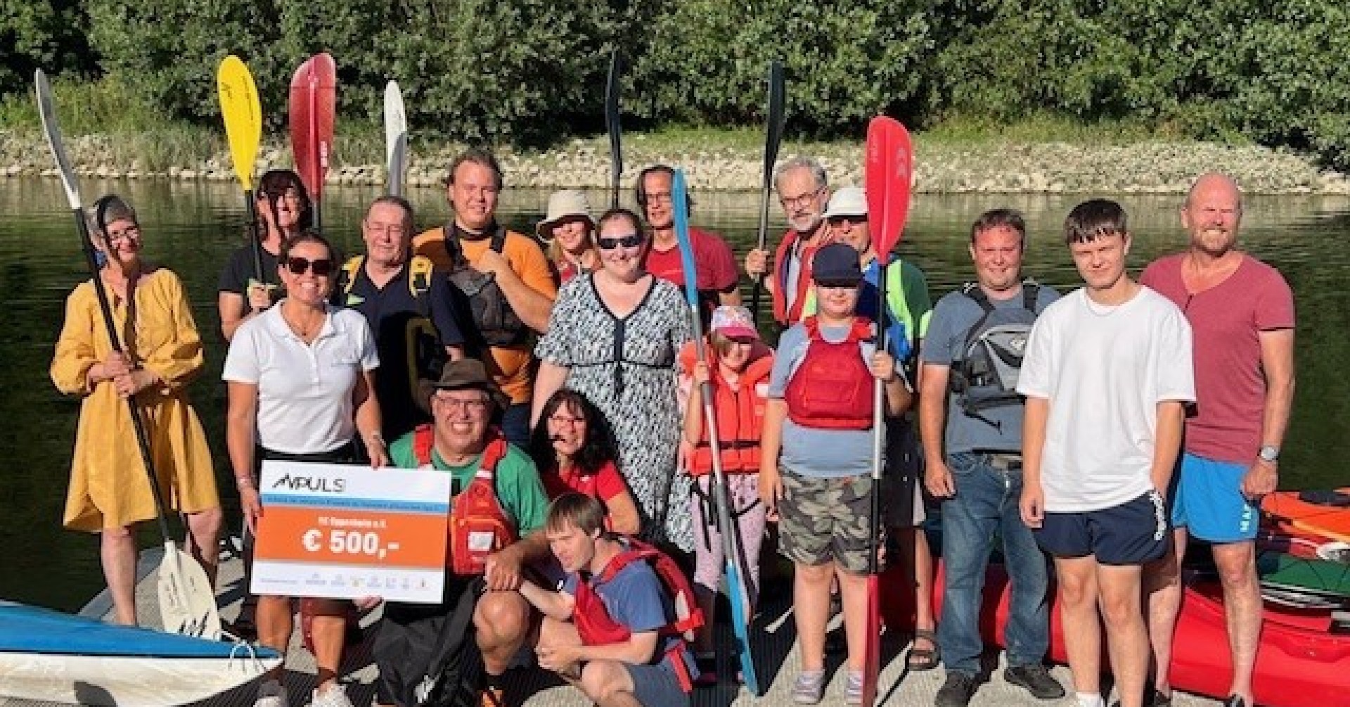 Gruppenfoto Kanufahren mit Scheckübergabe von APULS über 500 Euro am Flussufer Gruppe von Menschen beim Kanufahren, einige halten Paddel und einen großen Scheck mit der Aufschrift 'APULS' und 500 Euro, im Hintergrund ein Fluss und Bäume.