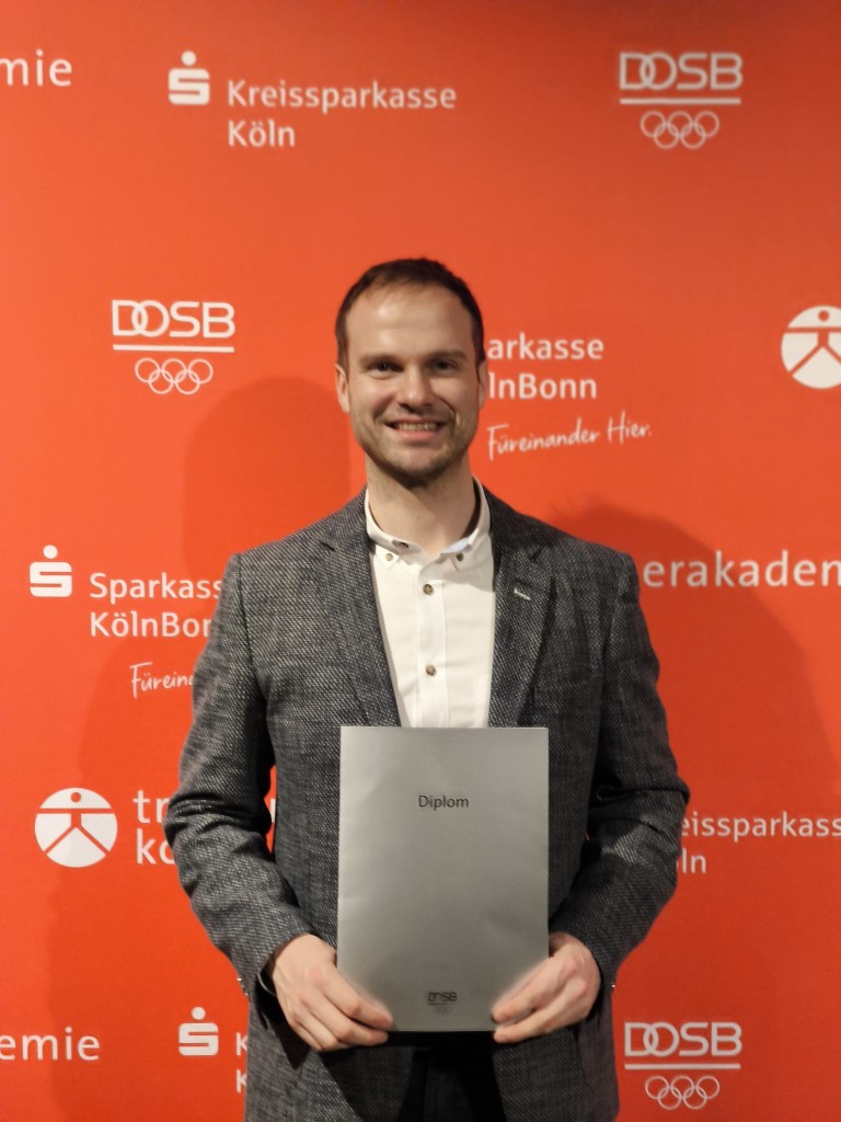 Diplomübergabe bei Sparkasse KölnBonn und DOSB – Preisverleihung oder Abschlussfeier Person mit Diplom vor Fotowand der Sparkasse KölnBonn und DOSB bei einer Preisverleihung oder Abschlussfeier