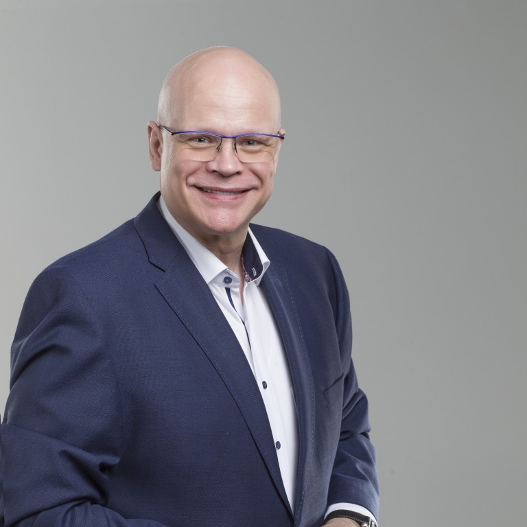Dr. Thomas Ellwanger Lächelnder Mann in blauem Anzug mit Brille vor grauem Hintergrund