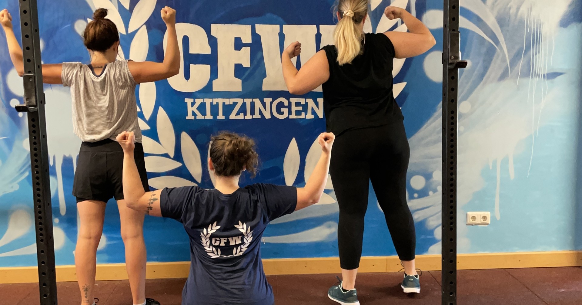 Drei Personen zeigen Muskeln vor einer CrossFit-Wand im Fitnessstudio.