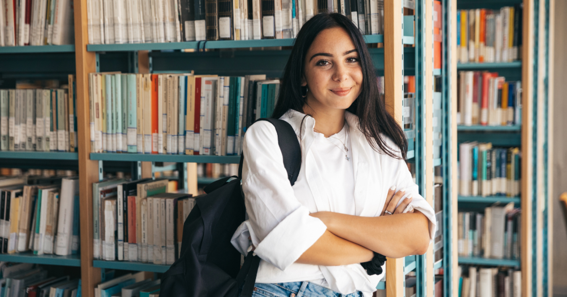 Studentin in weißem Hemd vor Bücherregal in der Bibliothek Studentin mit Rucksack steht vor Bücherregal in einer Bibliothek