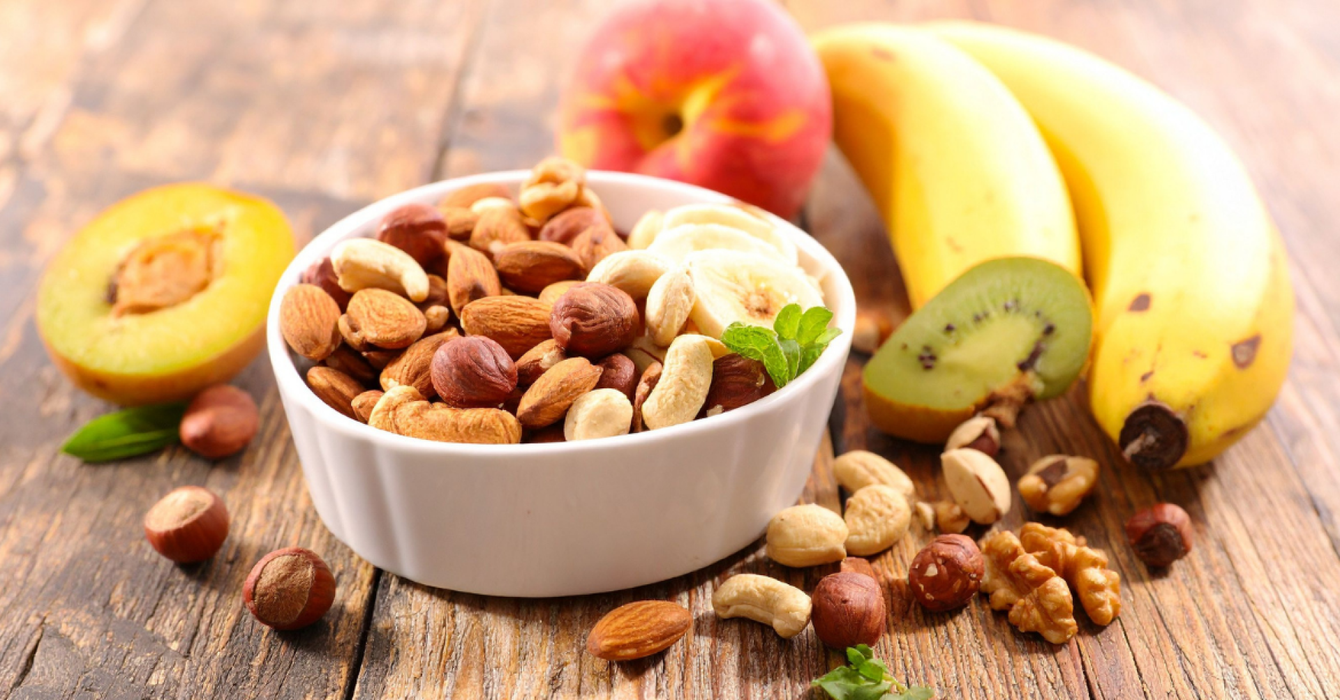 Schale mit Nüssen und Trockenfrüchten, daneben frische Banane, Kiwi und Pfirsich auf Holztisch
