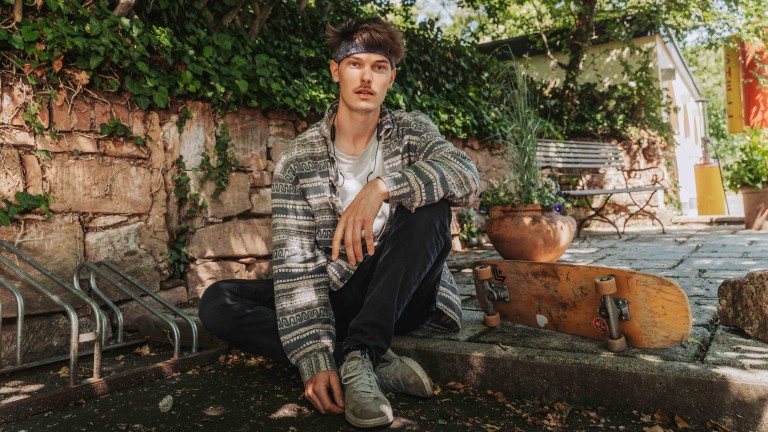 Junger Mann mit gemusterter Jacke und Skateboard im Garten, alea alternativ s 07