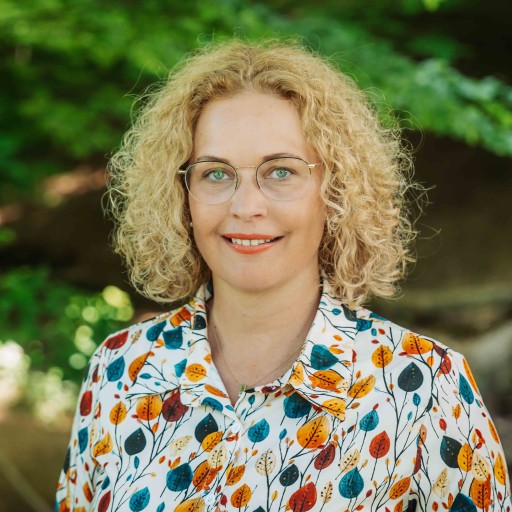 Alea Portrait – Frau mit lockigem Haar und bunter Bluse vor Naturhintergrund Alea Portrait einer Frau mit lockigem blondem Haar und bunter Bluse im Grünen