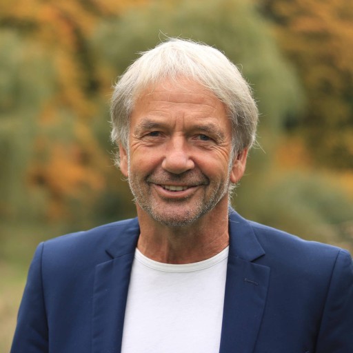 Mann mit grauen Haaren im blauen Sakko und weißem T-Shirt vor herbstlicher Naturkulisse