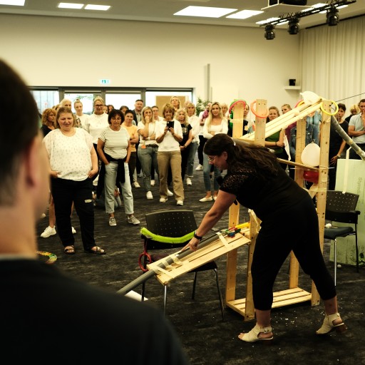 Gruppe von Menschen beobachtet eine Frau bei einer Teamaktivität mit einer selbstgebauten Kugelbahn in einem Konferenzraum