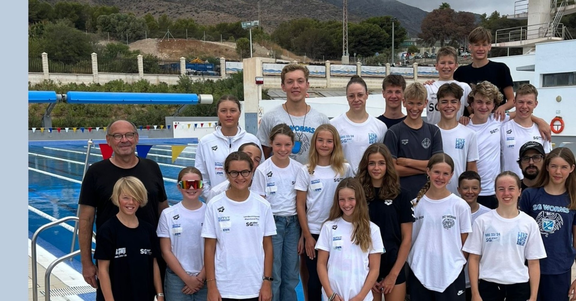 Gruppenfoto einer Schwimmgruppe am Beckenrand, aufgenommen im Freien mit Bergen im Hintergrund.