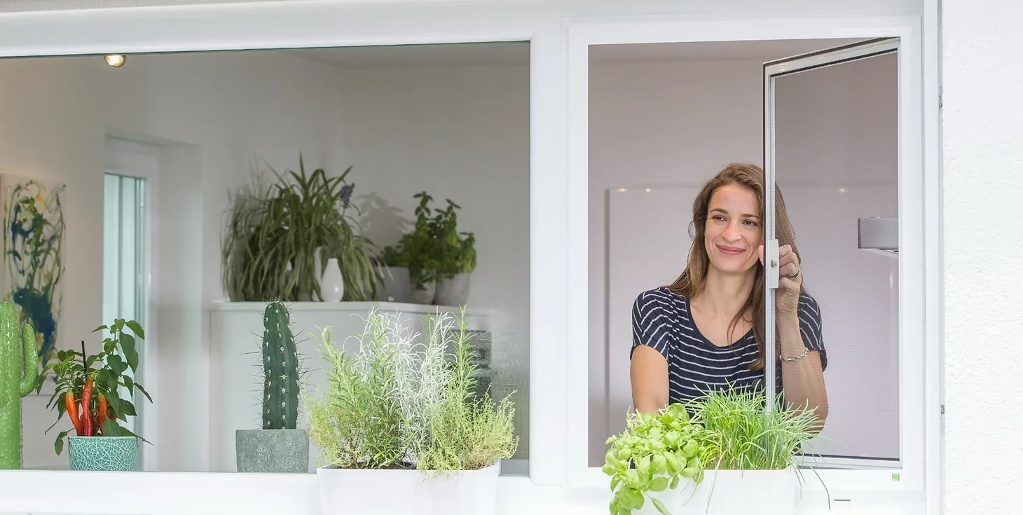 Lächelnde Frau am Fenster mit Kräutern und Pflanzen im Hintergrund