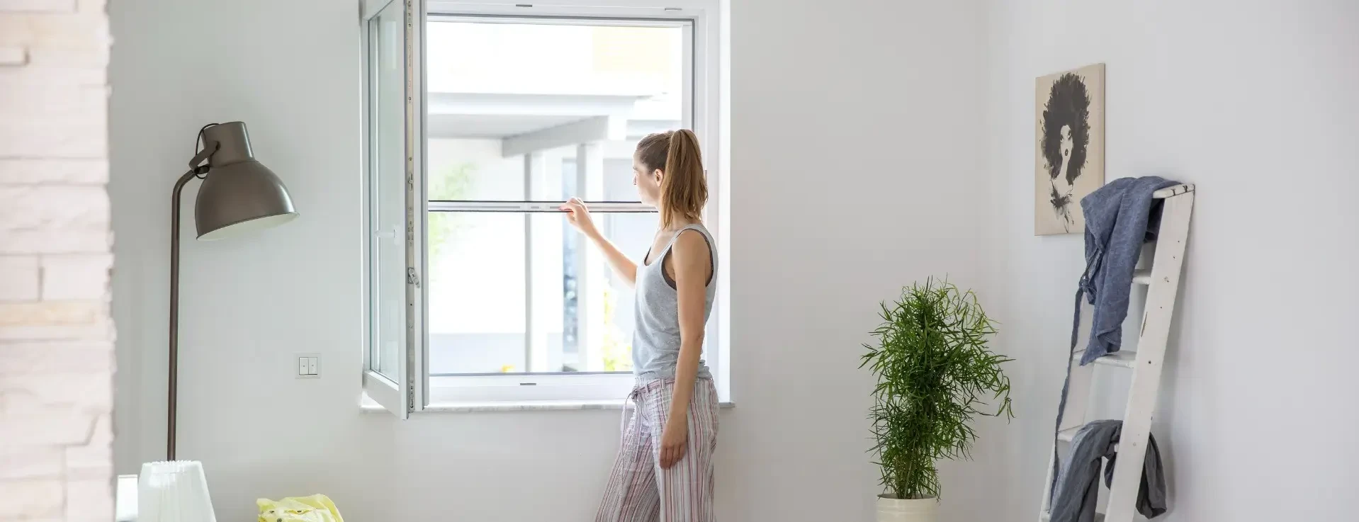 Frau im Pyjama öffnet ein Fenster mit Insektenschutz in einem hellen Raum