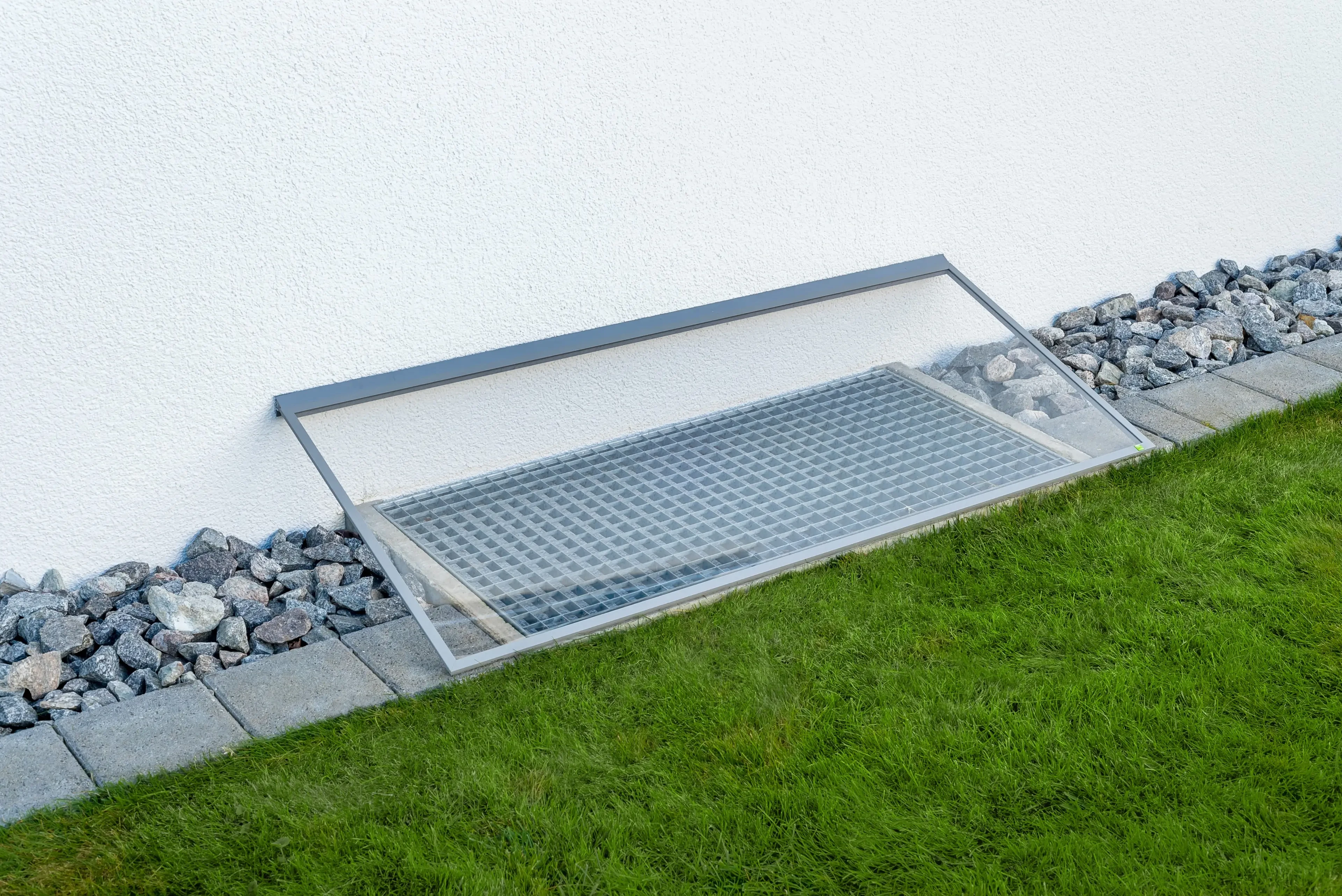 Transparente Lichtschachtabdeckung RESI im Garten wurde an einer weißen Hauswand mit angrenzendem Rasen und Kieselsteinen angebracht: Regenschutz und Belüftung des ursprünglichen Lichtschachts