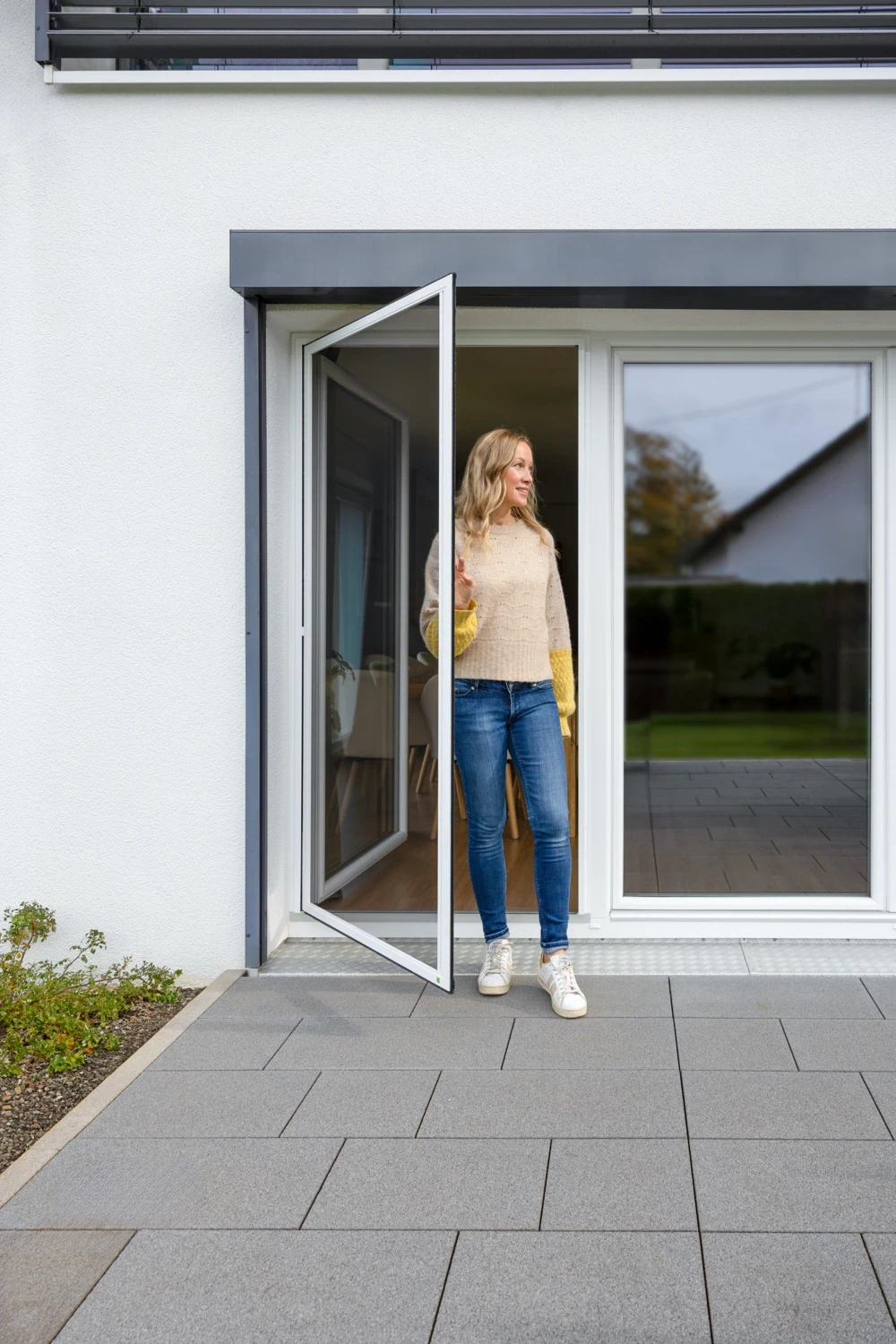 Frau in Jeans und Pullover tritt durch eine geöffnete Fenstertür auf eine Terrasse