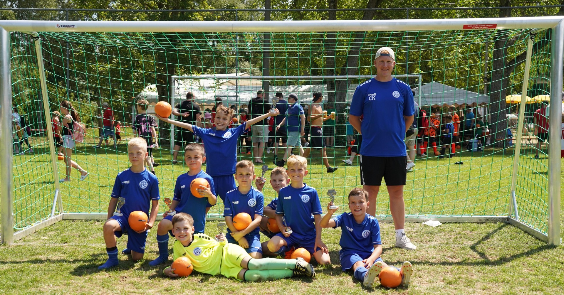 Junges Fußballteam posiert mit ihrem Trainer vor einem Tor auf einem Sportplatz, halten Pokale und Bälle.