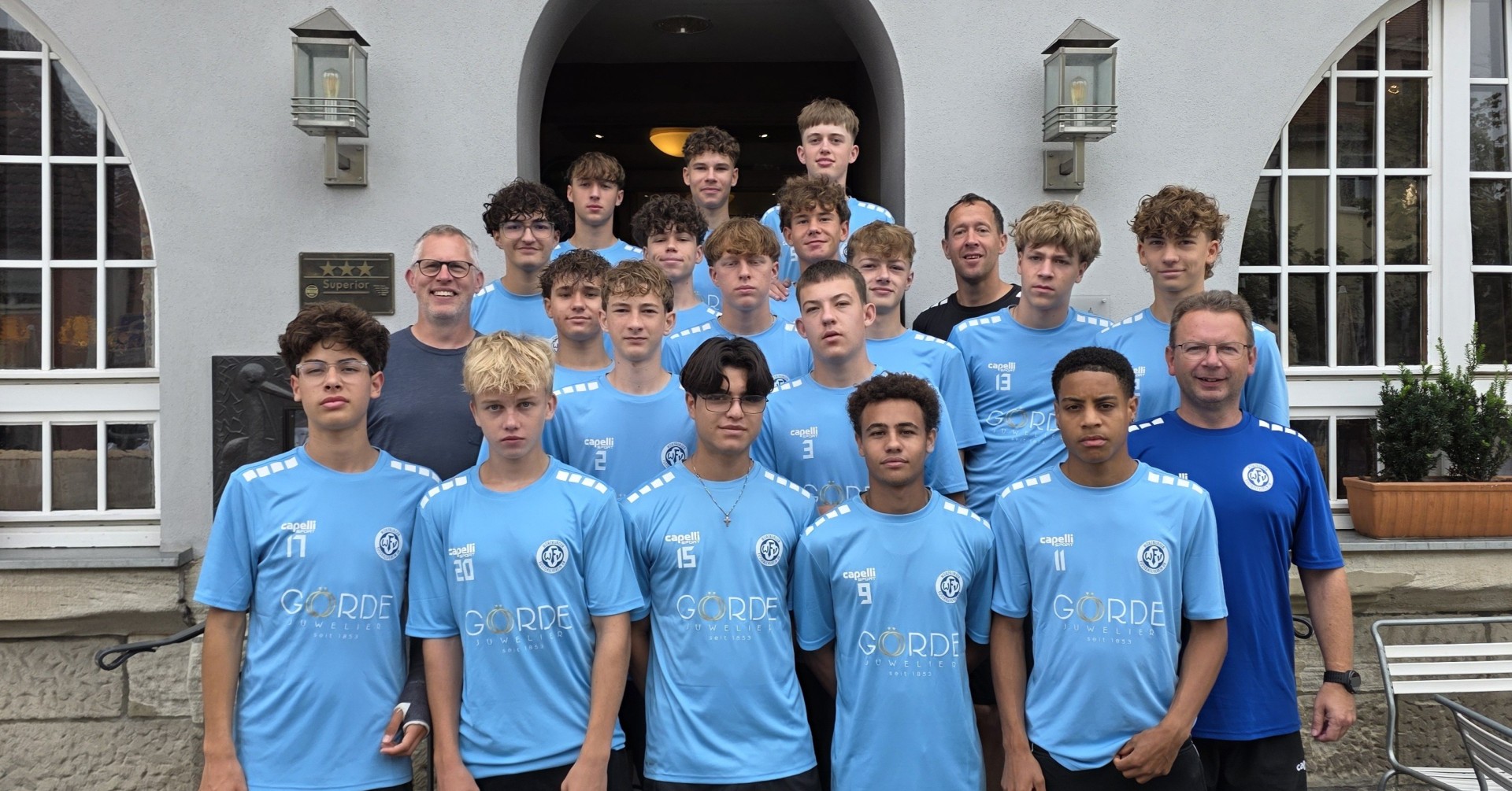 Jugend-Fußballmannschaft in hellblauen Trikots mit Sponsorenlogo Görde vor einem Hoteleingang
