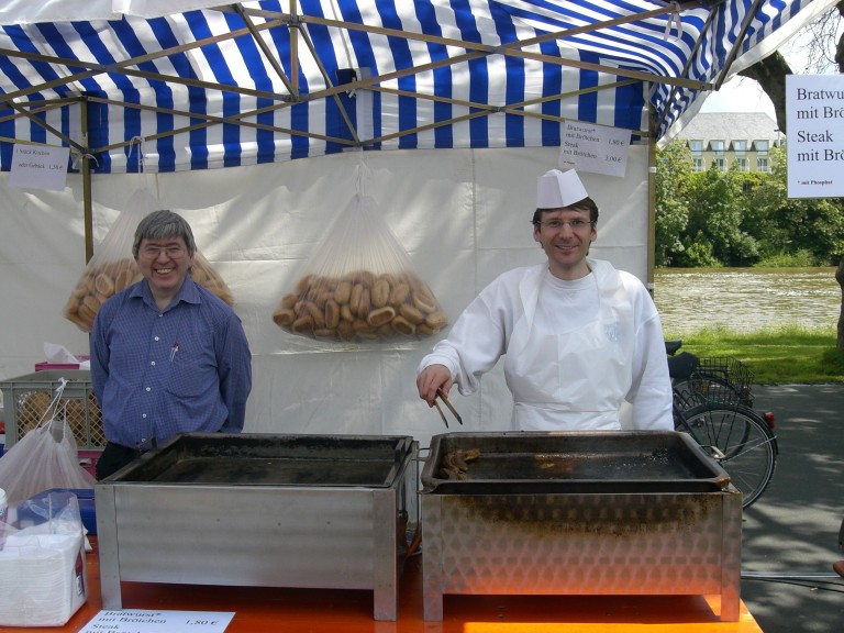 Hartmut und Roland am Grill auf dem Viehmarktplatz – Grillstand mit Bratwürsten und Brötchen Hartmut und Roland am Grill auf dem Viehmarktplatz bei einer Grillveranstaltung, Bratwurststand mit Brötchen unter blau-weißem Pavillon